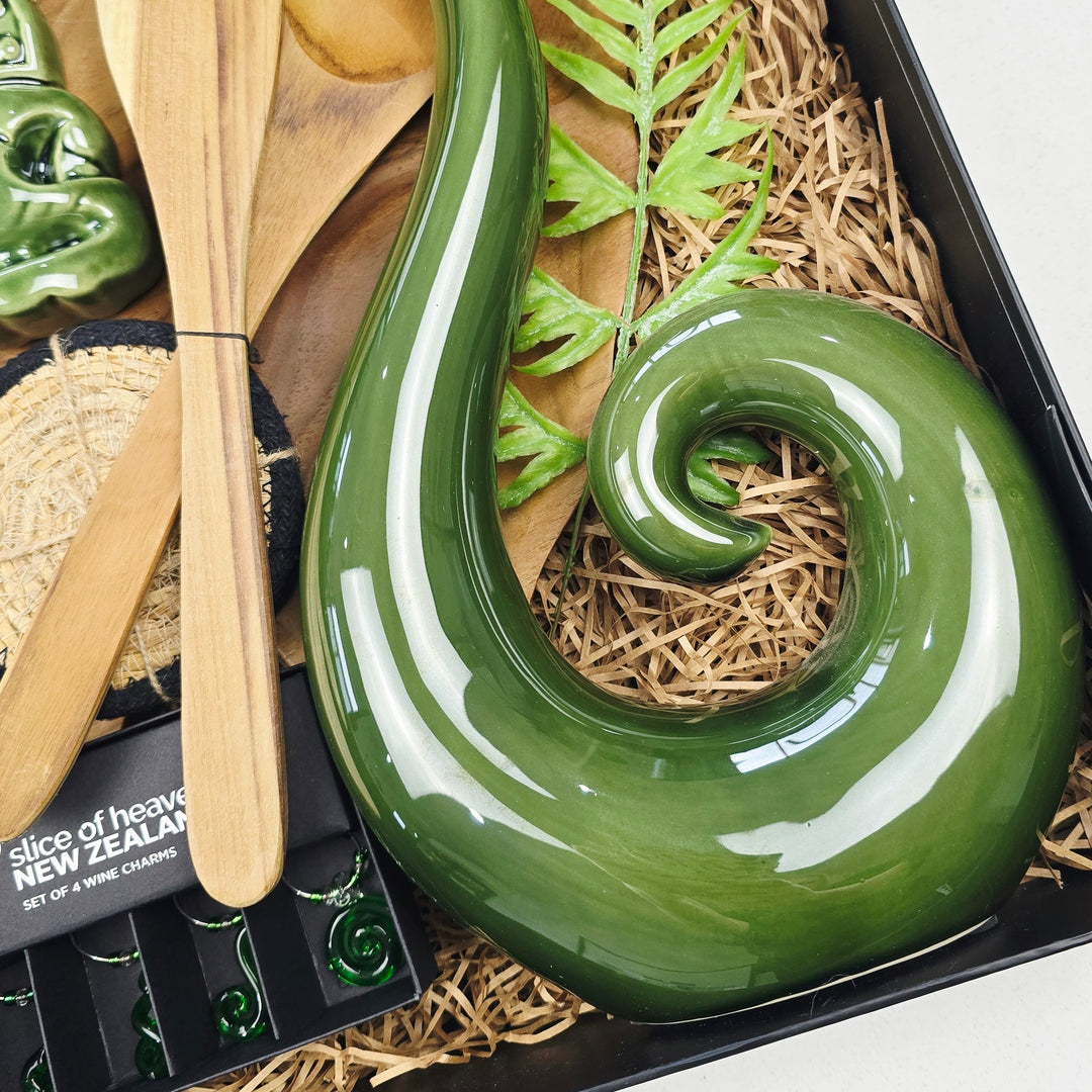 A green ceramic New Zealand tikanga carving presented in a black gift box with wooden utensils, a small green bowl, and a fern leaf on a light background.