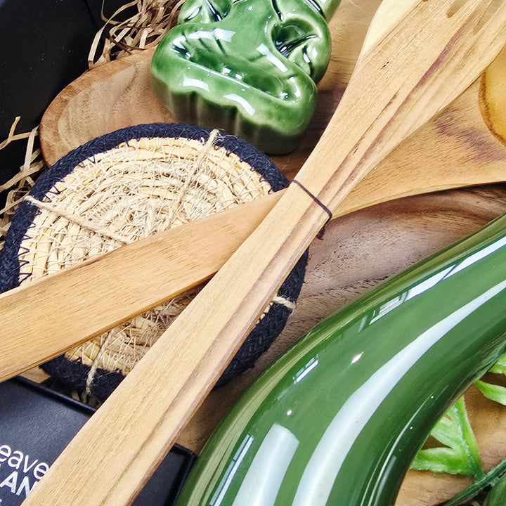 A close-up of a gift set containing a glossy green ceramic tiki mug, light natural bamboo utensils, a carved green ceramic tiki head, and a woven mat inside a black presentation box.