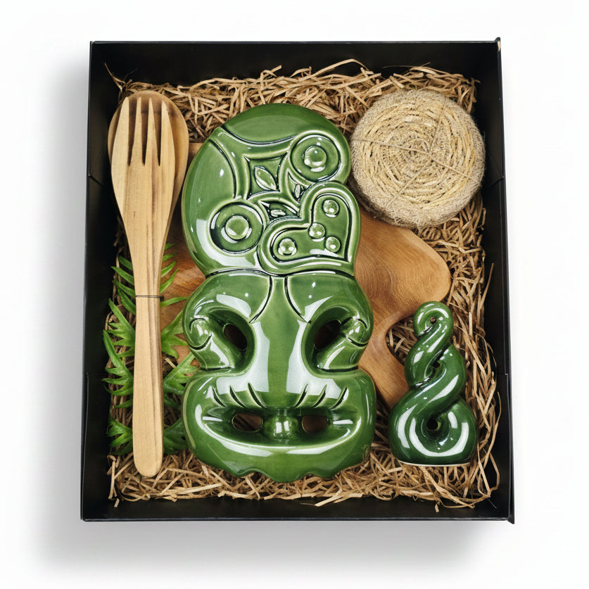 A green ceramic Maori-style carving set displayed inside a black gift box with natural wood and straw filler. The set includes a large carved figure, a smaller matching ornament, a light wood spoon, and a round woven fiber piece.