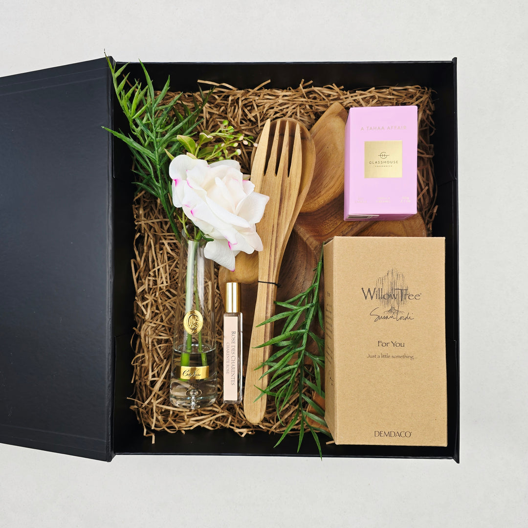 A curated gift box set from Dementia Co, featuring a black presentation box lined with brown paper shred, containing a wooden spoon and spatula, a small glass perfume bottle, a white rose, green sprigs, a pink box, and a kraft paper box labeled Willy's Bee Sugar Rush.