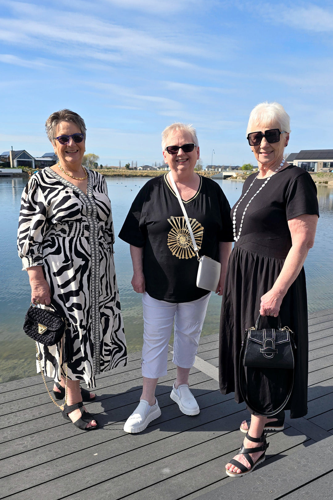 Three women posing together on a wooden dock by the water under a blue sky. They are dressed in black and white outfits, with one wearing a black dress, one in a black t-shirt with white pants, and one in a black and white zebra-patterned dress.