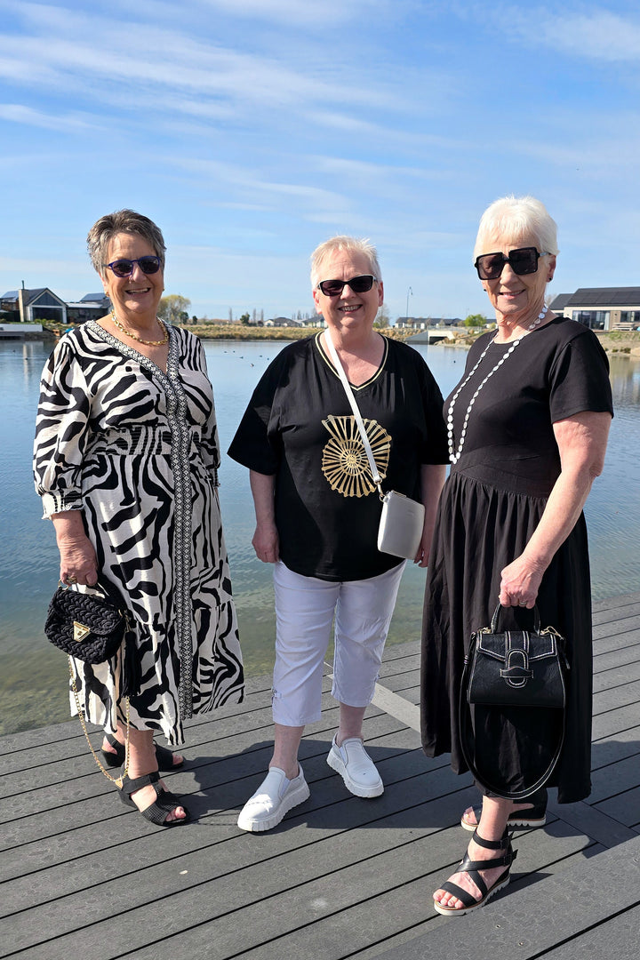 Three women posing together on a wooden dock by the water under a blue sky. They are dressed in black and white outfits, with one wearing a black dress, one in a black t-shirt with white pants, and one in a black and white zebra-patterned dress.