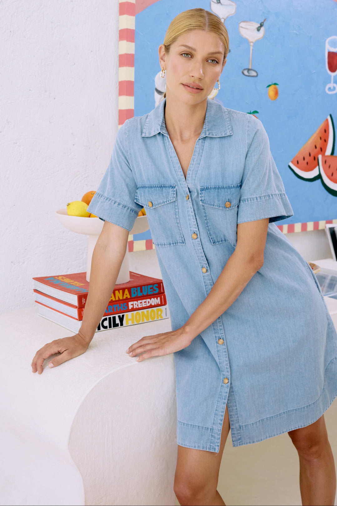 Woman wearing a light blue dress standing in front of a colorful wall with books and a watermelon illustration.