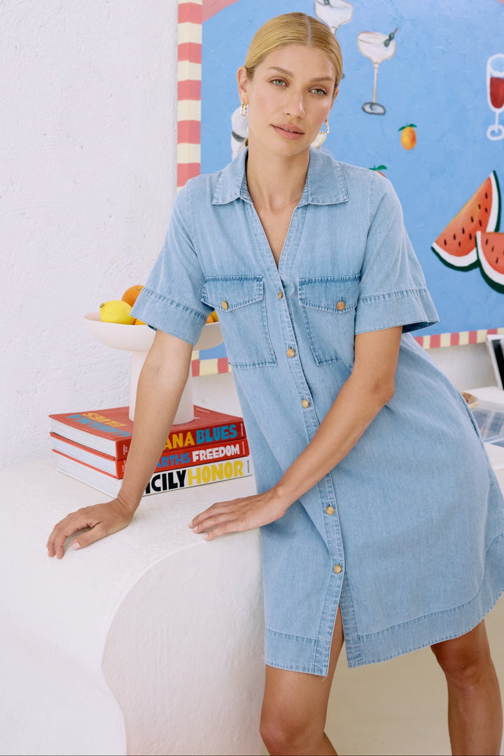 Woman wearing a light blue dress standing in front of a colorful wall with books and a watermelon illustration.
