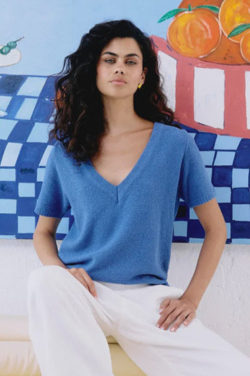Woman sitting on a couch wearing a blue top and white pants with a colorful mural in the background.