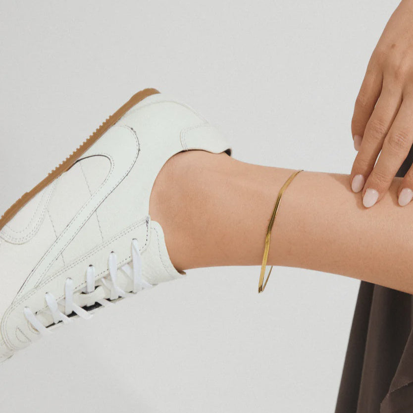 Person wearing a white sneaker with a gold bracelet on a plain background