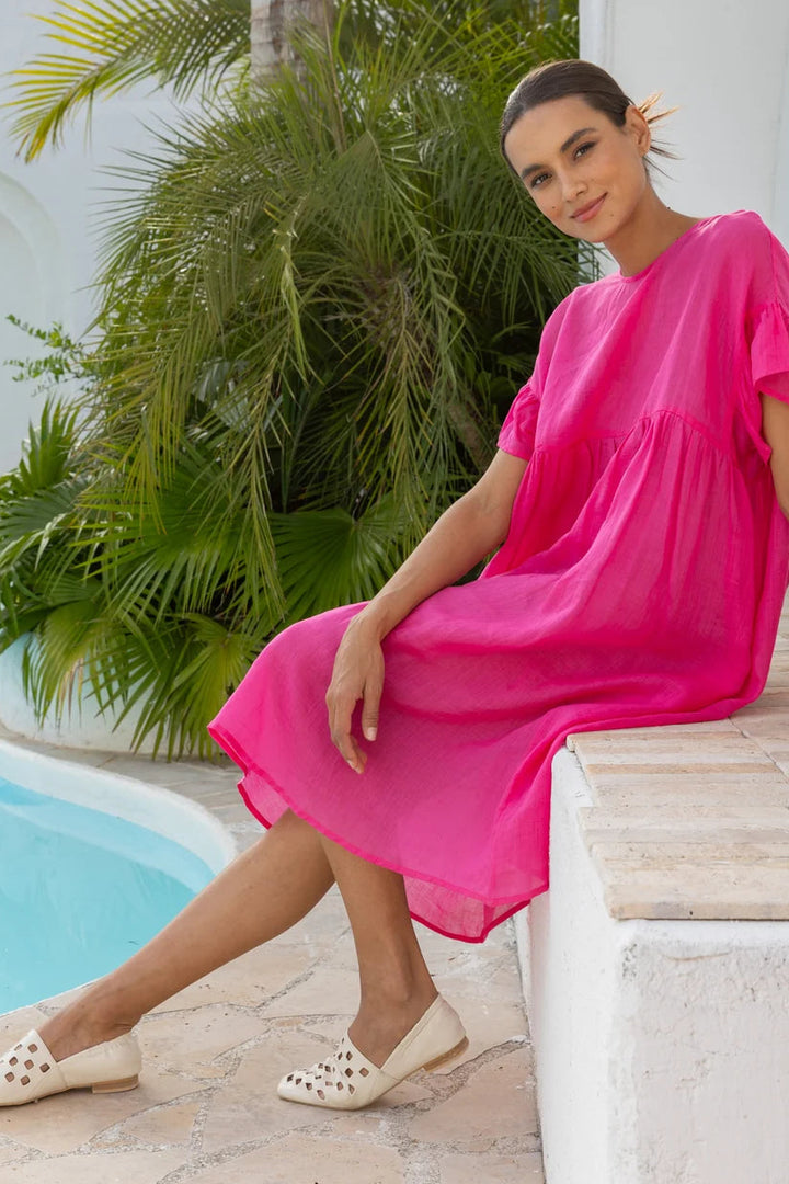 A woman wearing a bright pink tiered midi dress and beige studded flats sits on a white stone ledge next to a swimming pool with lush green palm foliage in the background.