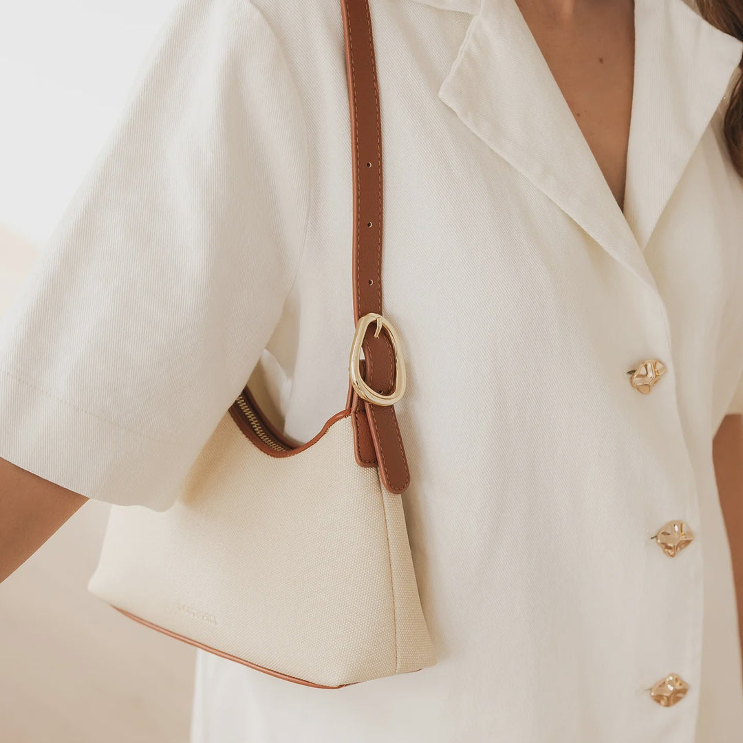 Close-up of a person wearing a white blazer with gold buttonswith a brown and cream canvas louenhide handbag.