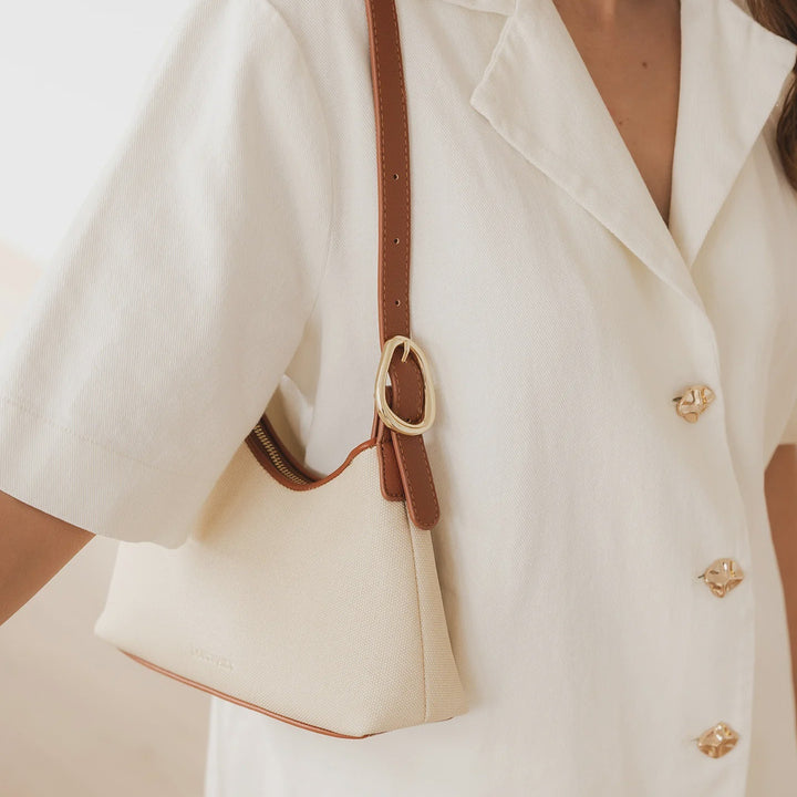Close-up of a person wearing a white blazer with gold buttonswith a brown and cream canvas louenhide handbag.