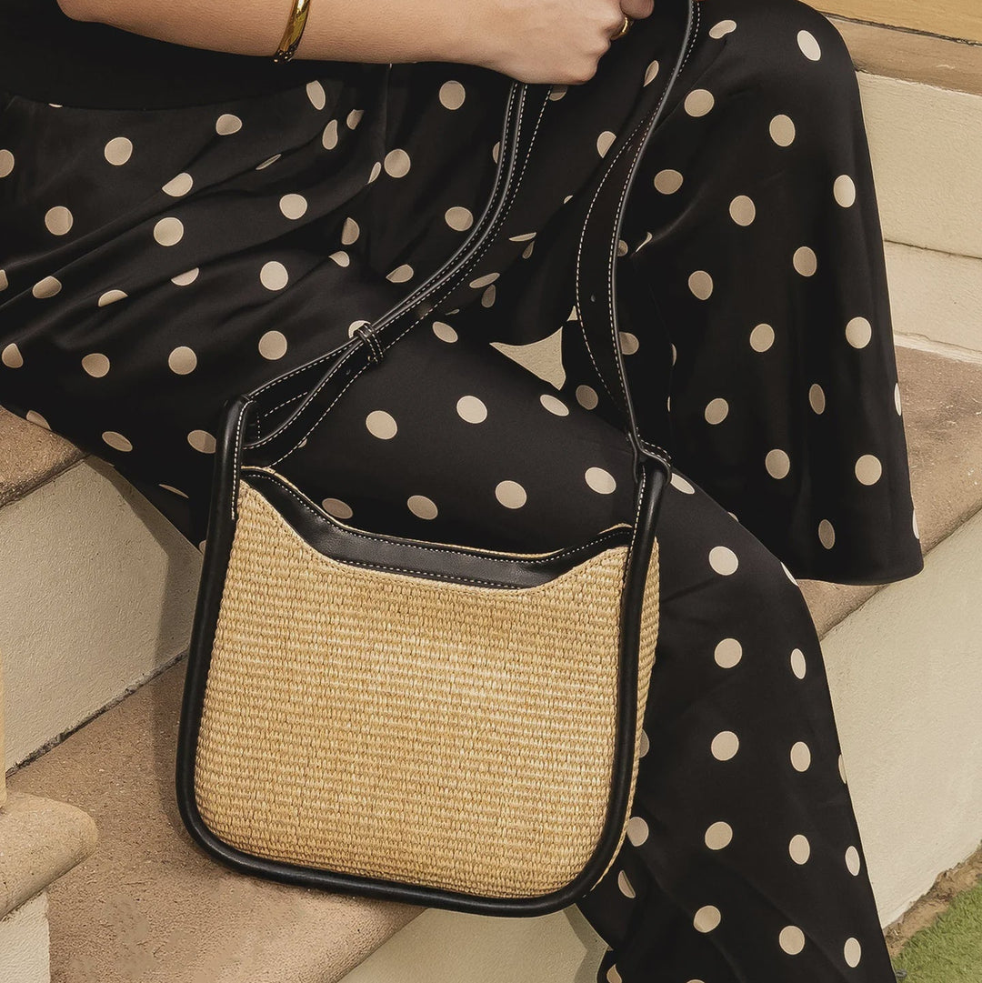 Person wearing a black dress with white polka dots holding a raffia louenhide summer handbag.