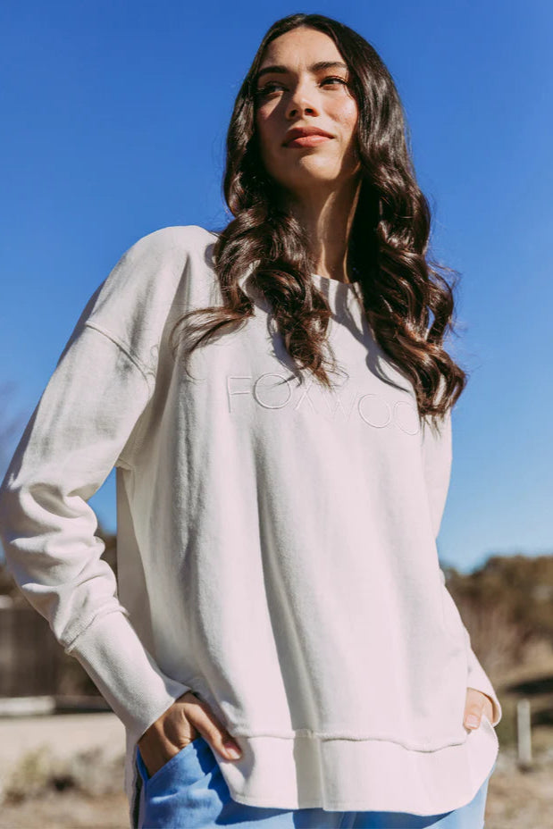 Woman wearing a light gray sweatshirt with a brand logo outdoors against a blue sky.