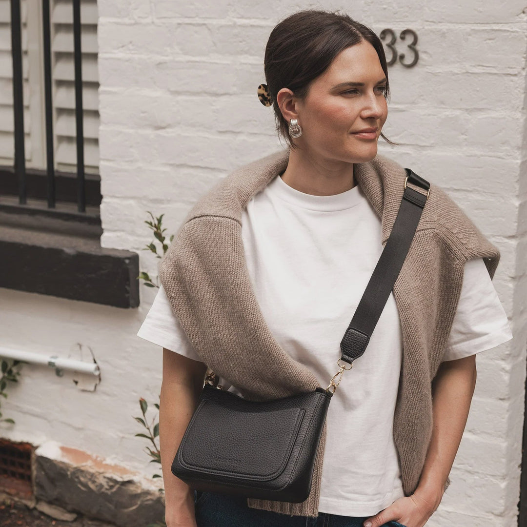 Woman wearing a beige shrug over a white shirt with a black crossbody bag, standing against a white brick wall.