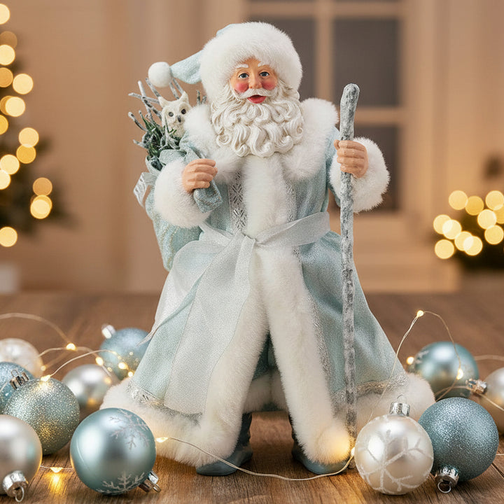 Decorative figure of a Possible Dreams collectable bearded santa figurine in a light blue robe with white fur trim, holding a staff and branches on in a christmas setting.
