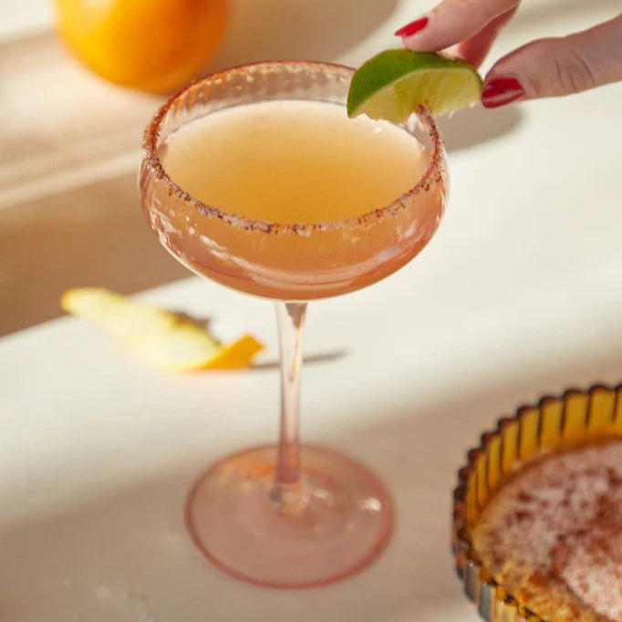 Cocktail with a lime wedge on a white surface with an orange and lemon in the background.
