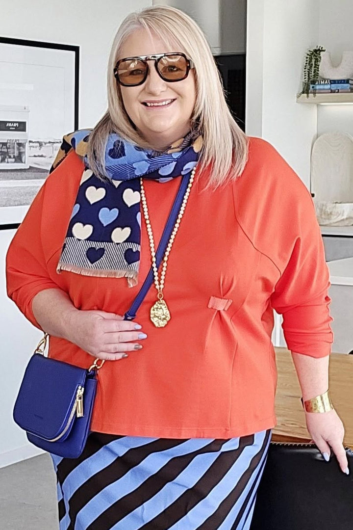 Woman in a bright orange top and blue striped skirt standing in a modern room with a desk and plants.