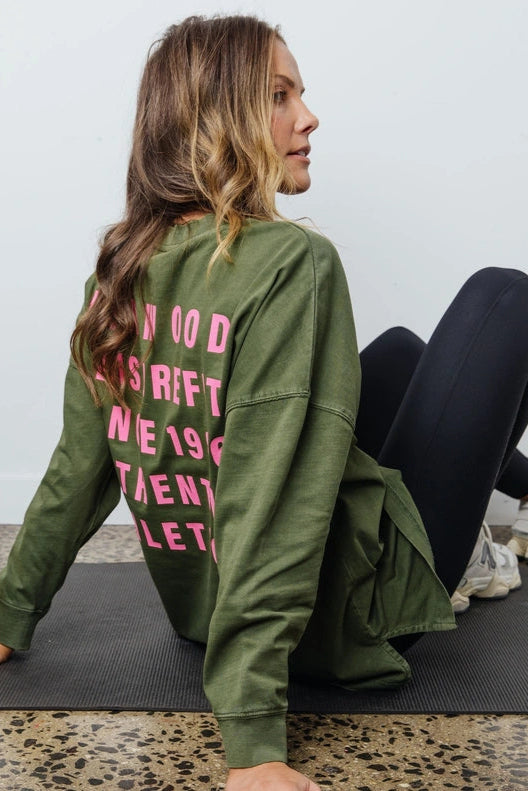 Woman sitting on a yoga mat wearing an olive green sweatshirt with pink text.