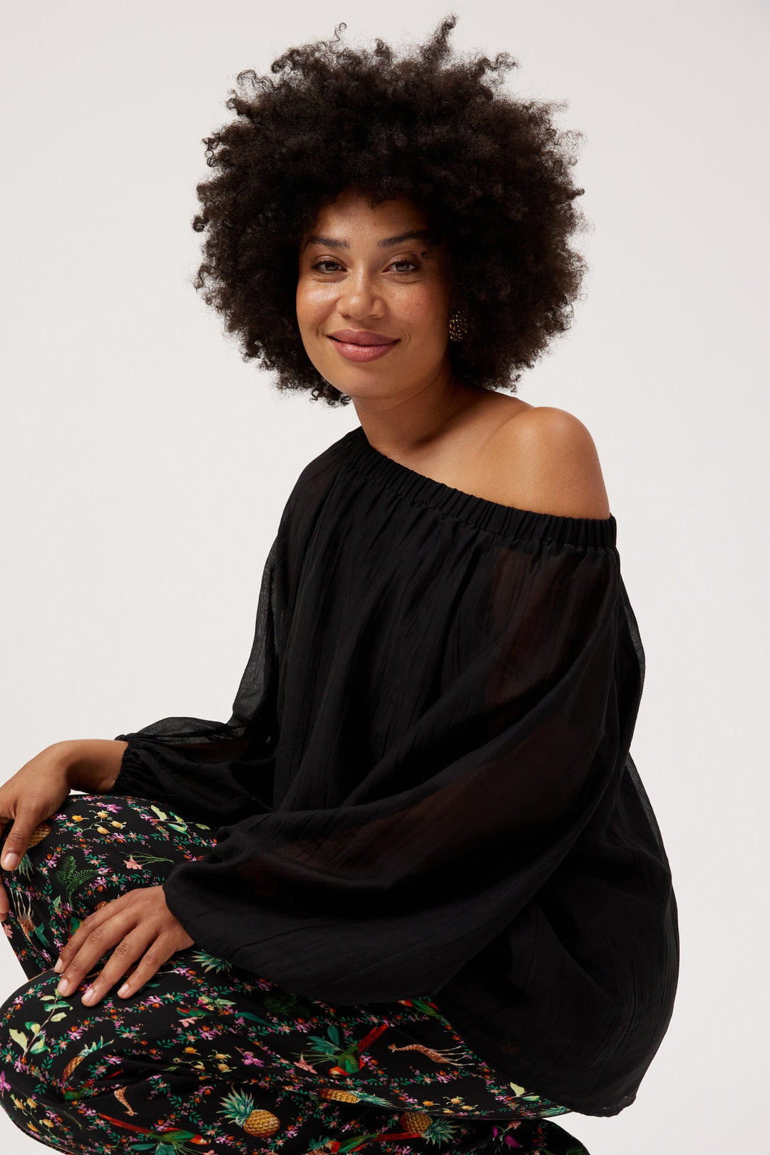 A woman with an afro hairstyle crouches in a studio setting, wearing a black off-the-shoulder sheer long-sleeve top and black wide-leg pants with a colorful floral and abstract print, paired with tan heeled sandals.