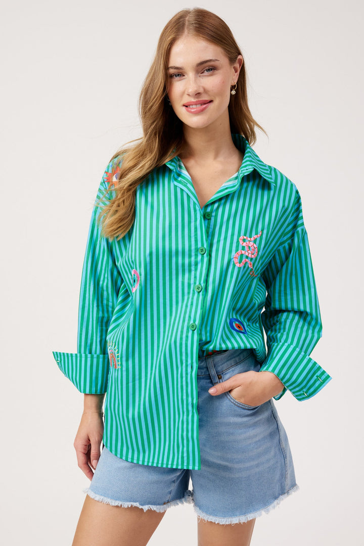 A woman modeling a green and white vertically striped button-up shirt with colorful floral embroidery on the chest, paired with light blue denim shorts, against a plain light gray studio background.