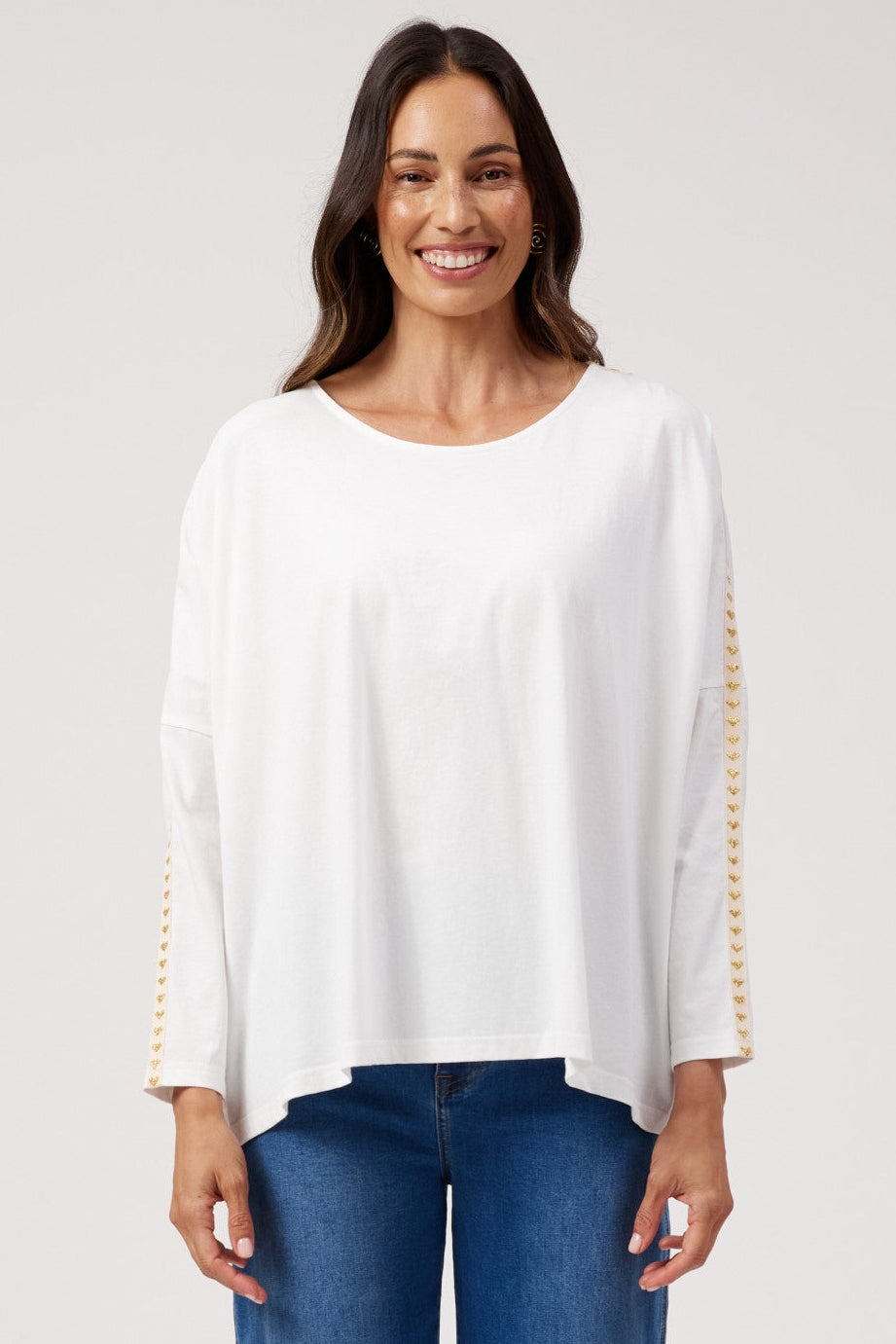 A woman modeling a white long-sleeve top with gold beaded trim on the sleeves, paired with blue wide-leg jeans, against a light gray studio background.