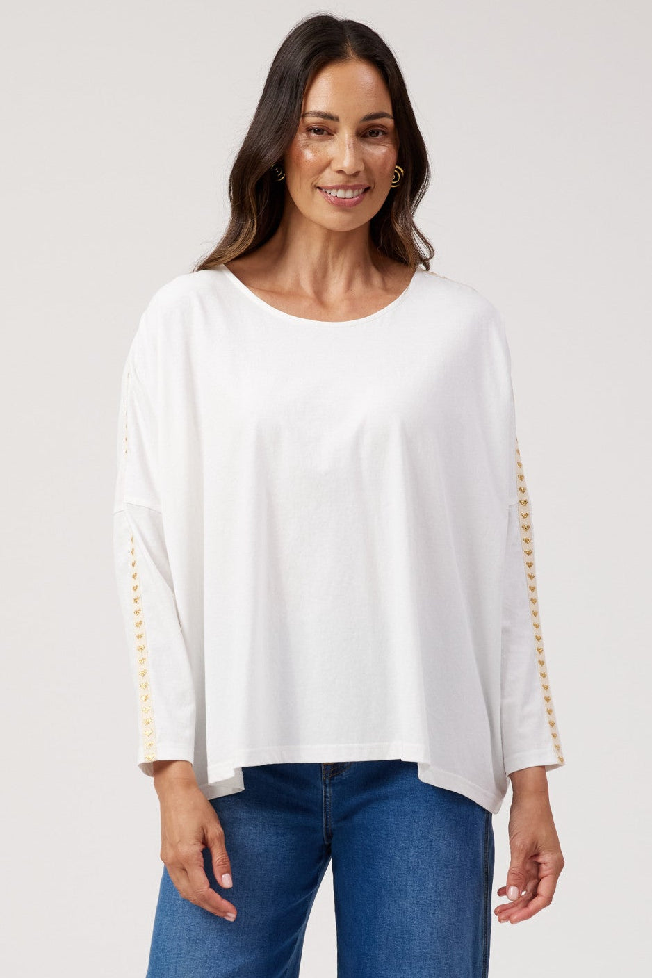 A woman modeling a white long-sleeve top with gold beaded trim on the cuffs, paired with wide-leg blue jeans and flat sandals, against a light gray studio background.