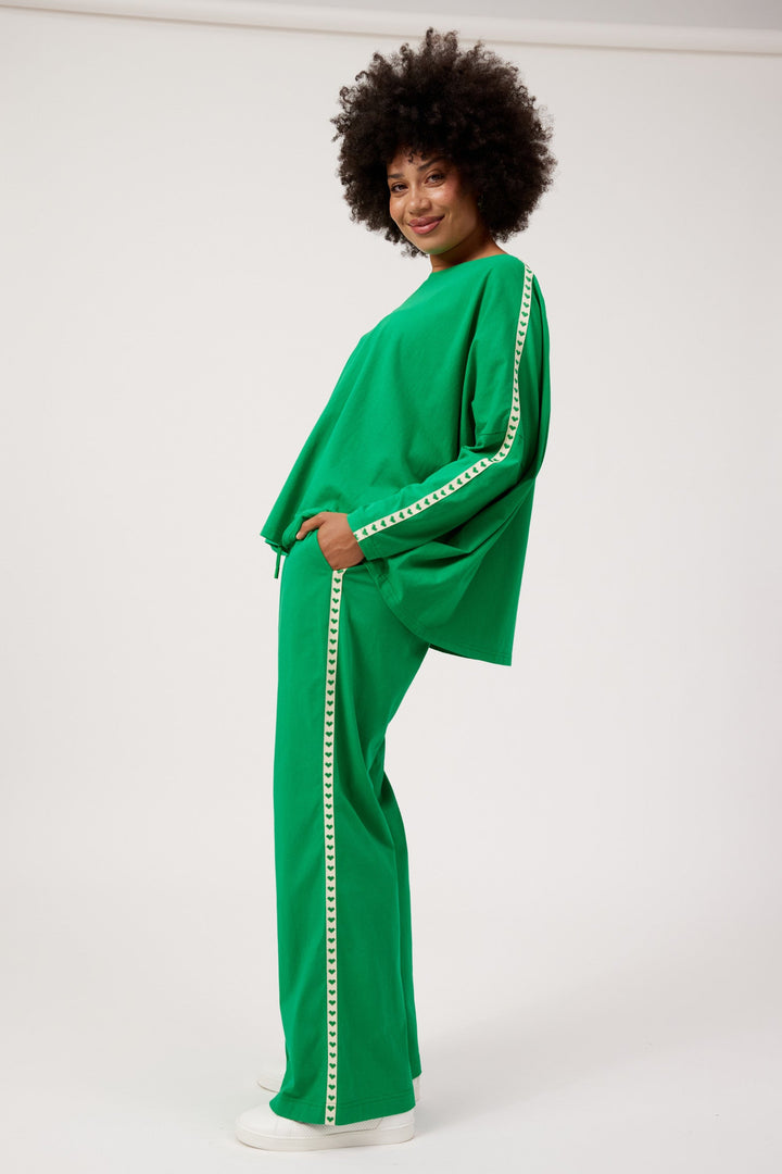 A woman with an afro hairstyle models a bright green two-piece lounge set featuring wide-leg pants and a matching long-sleeve top with white trim, set against a plain white studio background.