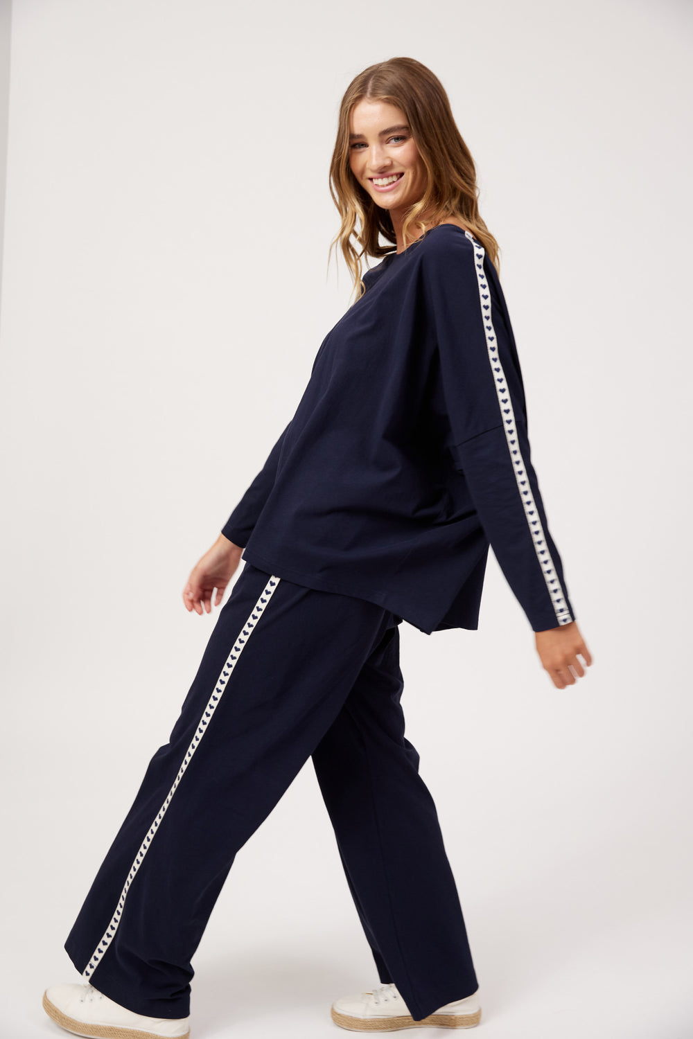A woman modeling a matching navy blue lounge set with white chain-link trim down the sleeves and pants, posed against a clean white studio background.