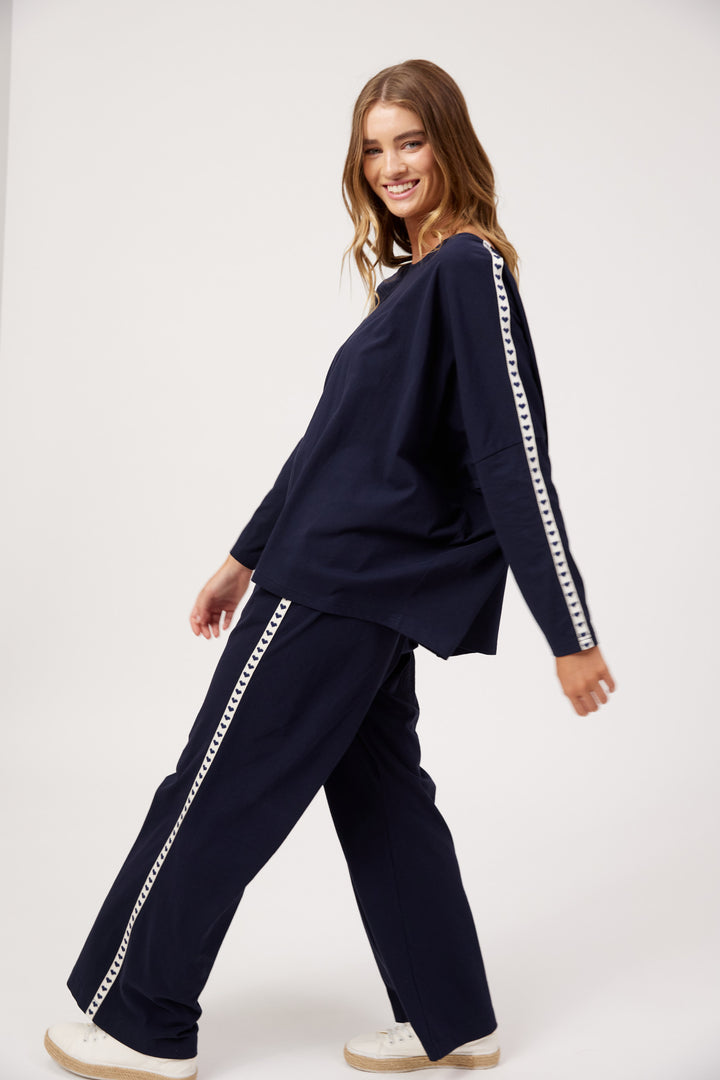A woman modeling a matching navy blue lounge set with white chain-link trim down the sleeves and pants, posed against a clean white studio background.