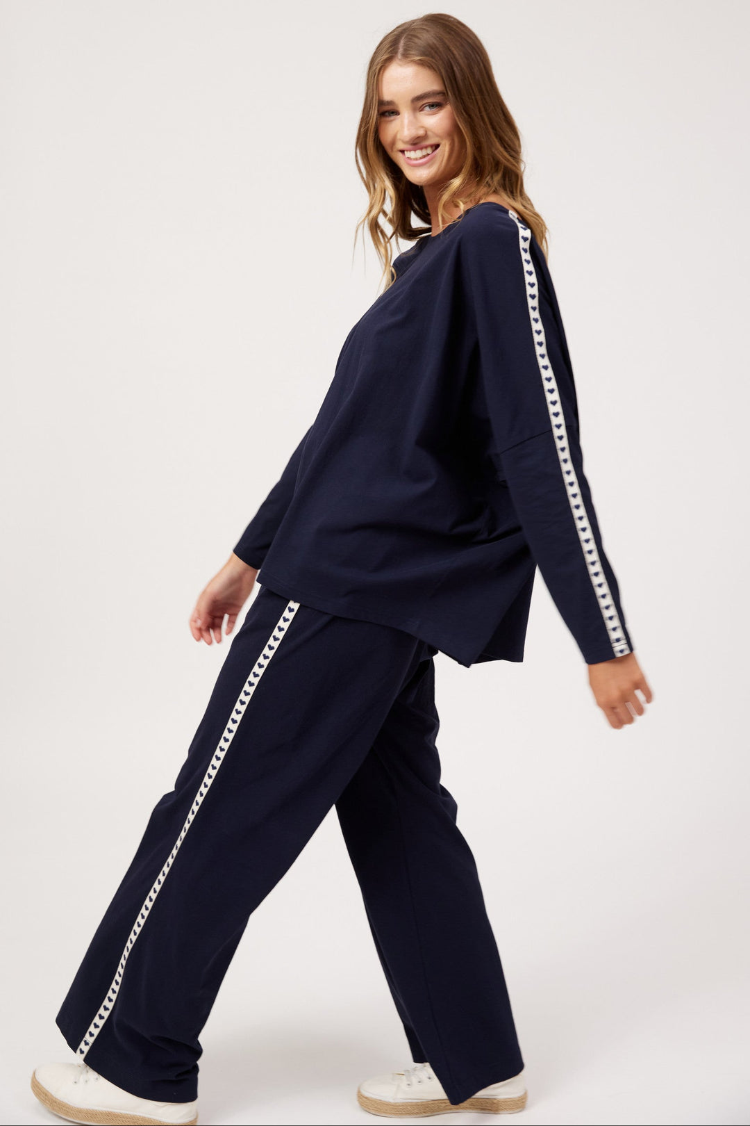 A woman modeling a matching navy blue lounge set with white chain-link trim down the sleeves and pants, posed against a clean white studio background.