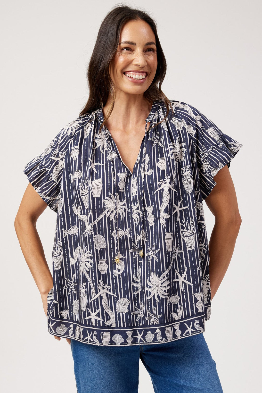 A woman modeling a short-sleeve navy blue blouse with a white palm tree and shell pattern, paired with blue jeans, against a plain light gray background.