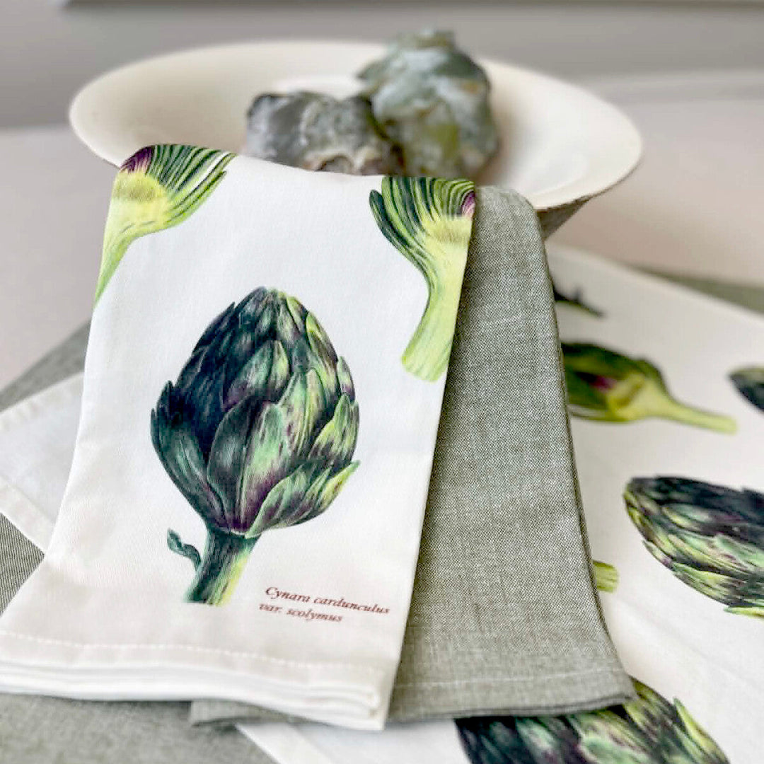 A folded white kitchen towel with a detailed artichoke illustration and small German text, layered over a gray linen napkin, with more matching towels and a white bowl in the background.