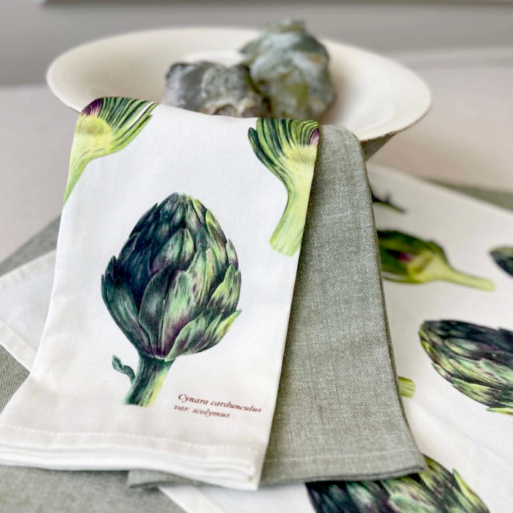 A folded white kitchen towel with a detailed artichoke illustration and small German text, layered over a gray linen napkin, with more matching towels and a white bowl in the background.