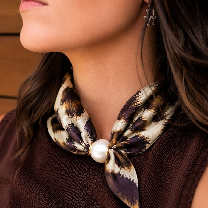 A close-up of a woman wearing a leopard-print silk neck scarf tied in a bow with a central white pearl, paired with a brown knit top.
