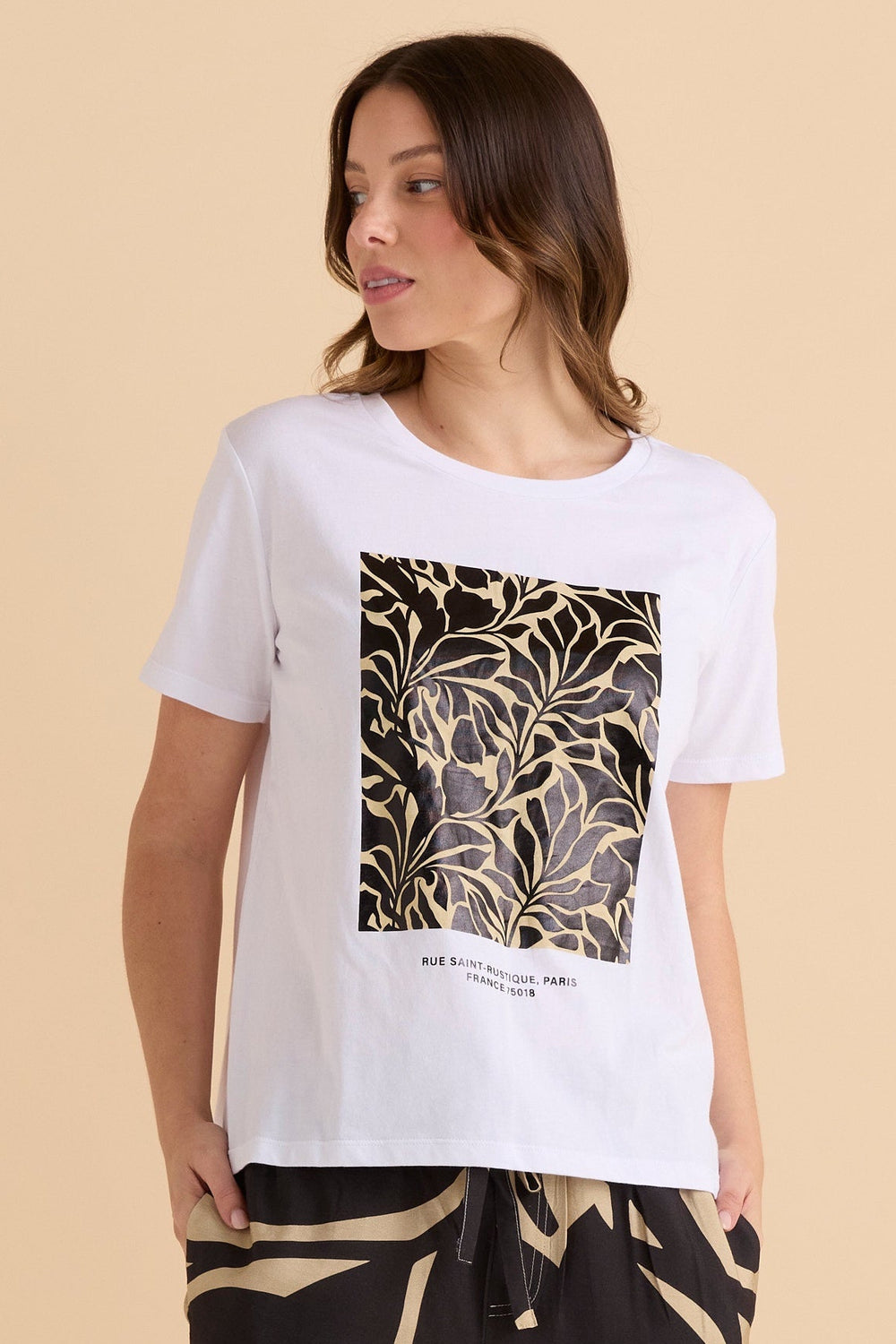 Woman wearing a white Btty Basics Ellia t-shirt with a black and gold leaf pattern on a beige background