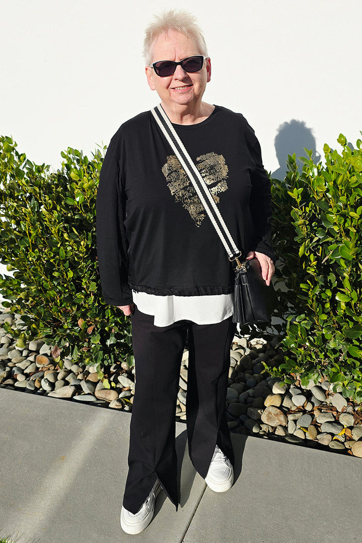 An older woman stands outdoors on a sidewalk wearing a black long-sleeve top with a graphic print, black pants, white sneakers, and a black shoulder bag with white straps. She is positioned in front of green hedges and a white wall.