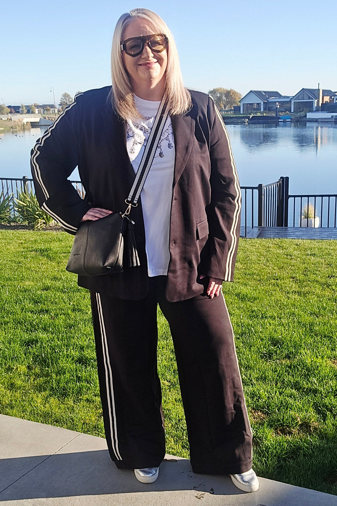 Person standing outdoors by a waterfront with green grass and a clear sky. Lady wearing a black & white stripe pants and blazer with a white tee.