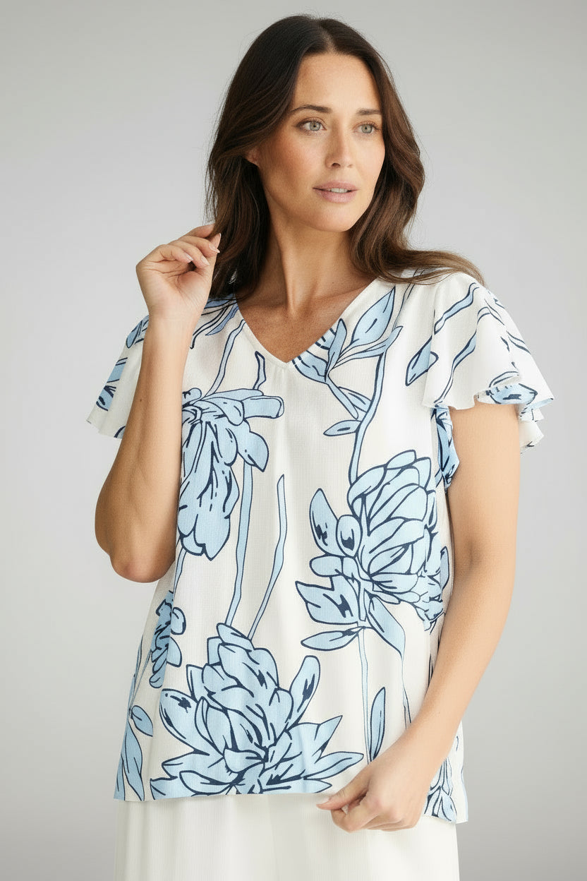 Woman wearing a blue floral top brave & true jemma and white pants on a white background