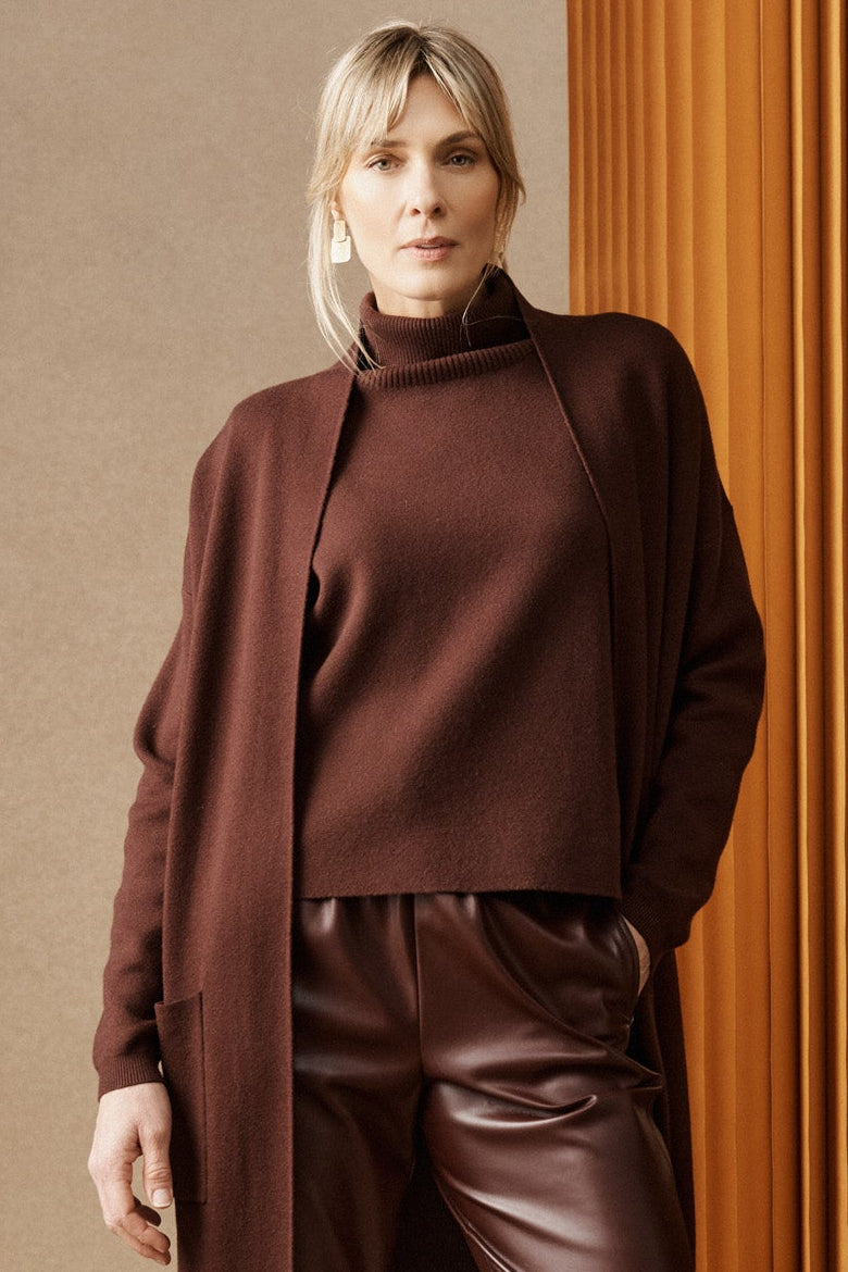 Two women standing against a textured wall, one in a brown outfit and the other in a white blouse with a brown skirt.