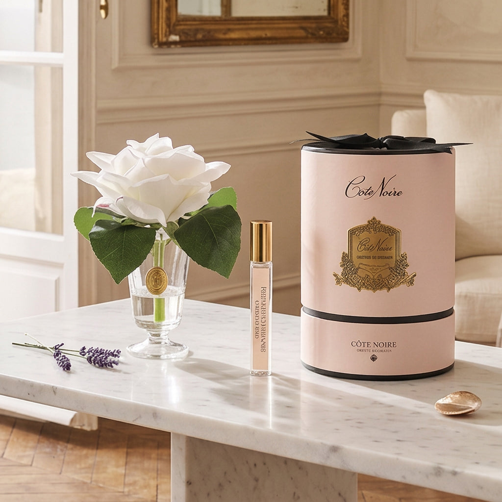 A gift set featuring a single white rose in a clear glass vase, a small perfume sample vial with a gold cap, and a tall pale pink cylindrical box with black ribbon and gold branding for Cote Noire.