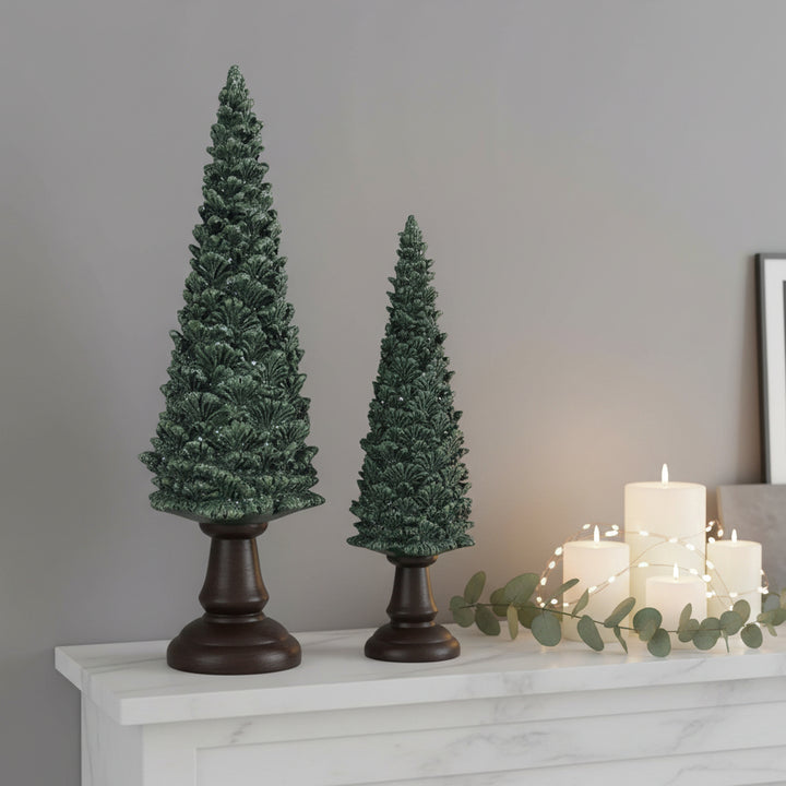 Two small artificial Christmas trees with dark green textured foliage and dark brown pedestal bases, shown on a plain white background.