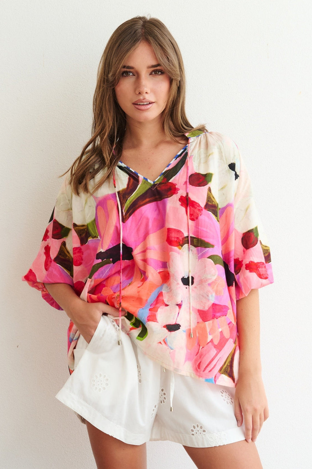 Woman wearing a colorful floral blouse and white shorts standing against a white wall.