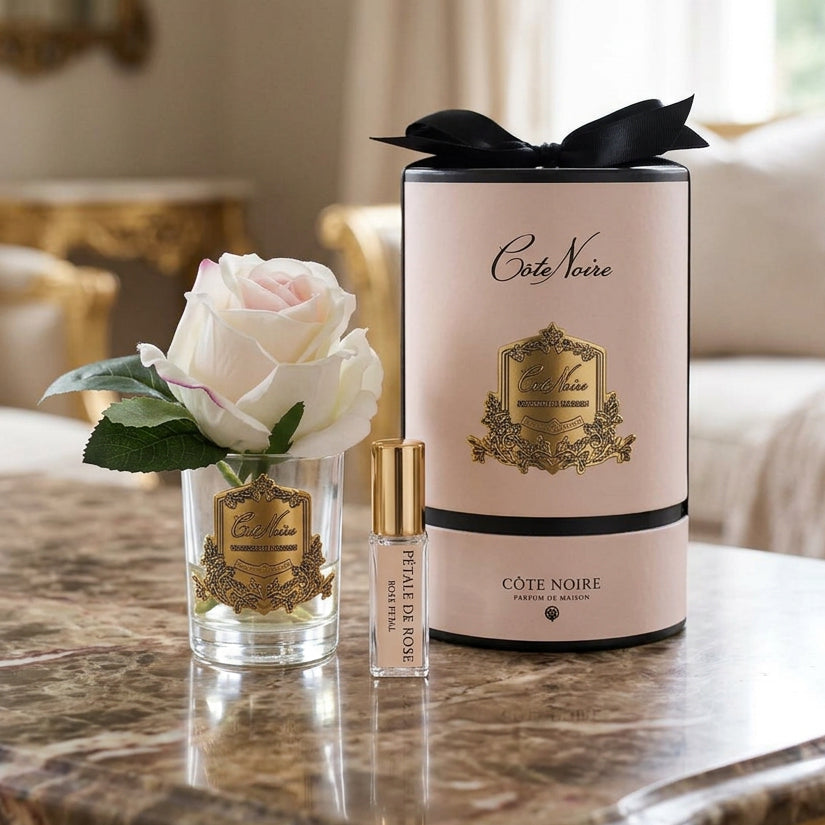 Cote Vierge fragrance set featuring a clear glass diffuser vase with a white rose, a small clear spray bottle with a gold cap, and a tall pale pink cylindrical box with black ribbon and gold crest detailing.