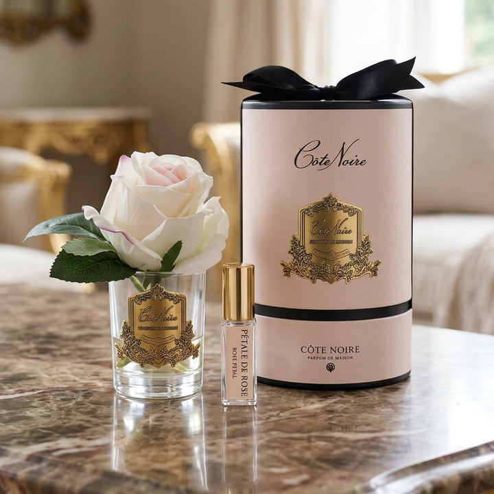 Cote Vierge fragrance set featuring a clear glass diffuser vase with a white rose, a small clear spray bottle with a gold cap, and a tall pale pink cylindrical box with black ribbon and gold crest detailing.
