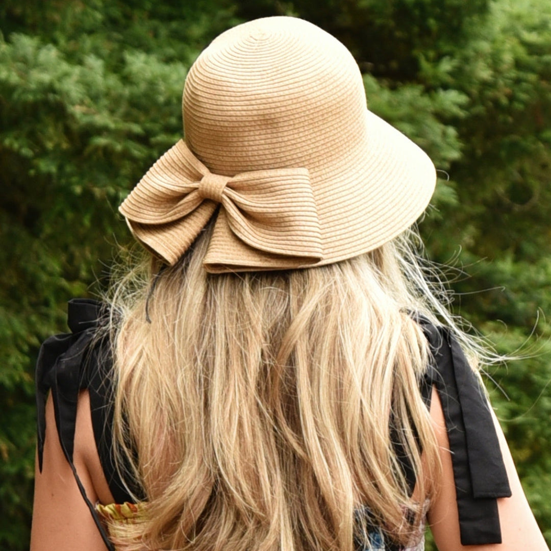 Person wearing a beige hat with a large bow, standing outdoors with greenery in the background.