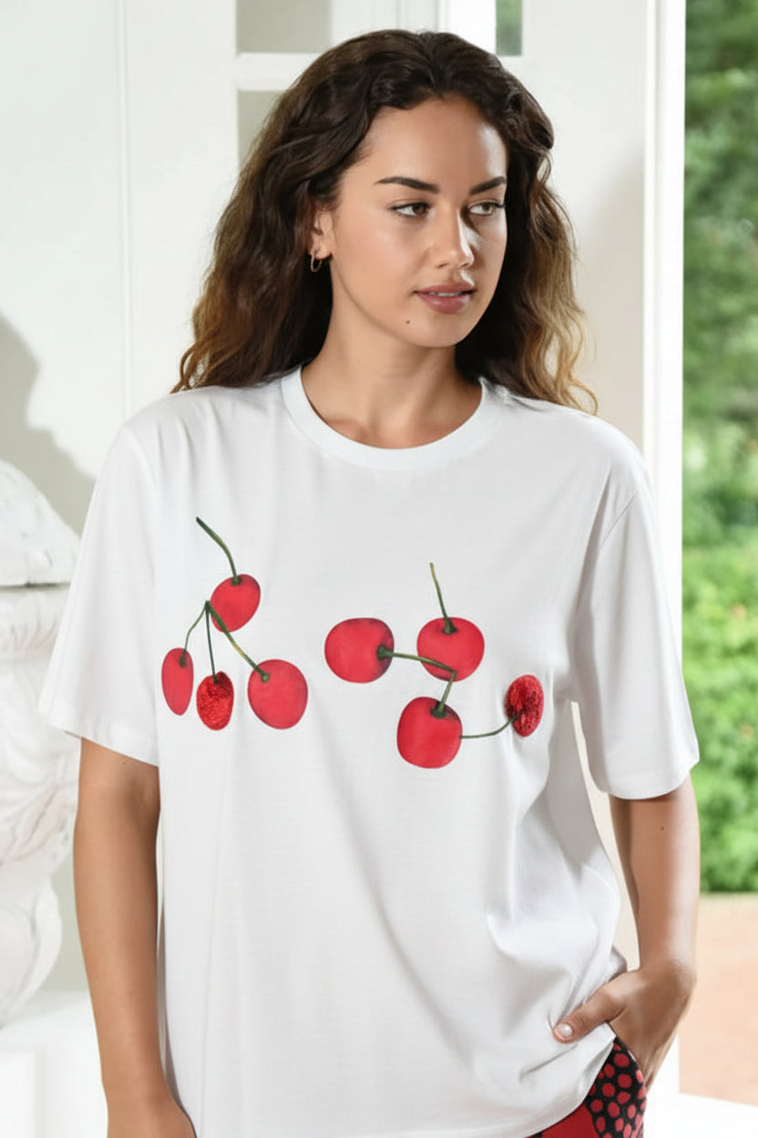 A woman wearing a coral-red short-sleeve t-shirt with a cherry graphic print on the front, paired with a floral-patterned skirt, standing in a bright indoor setting.
