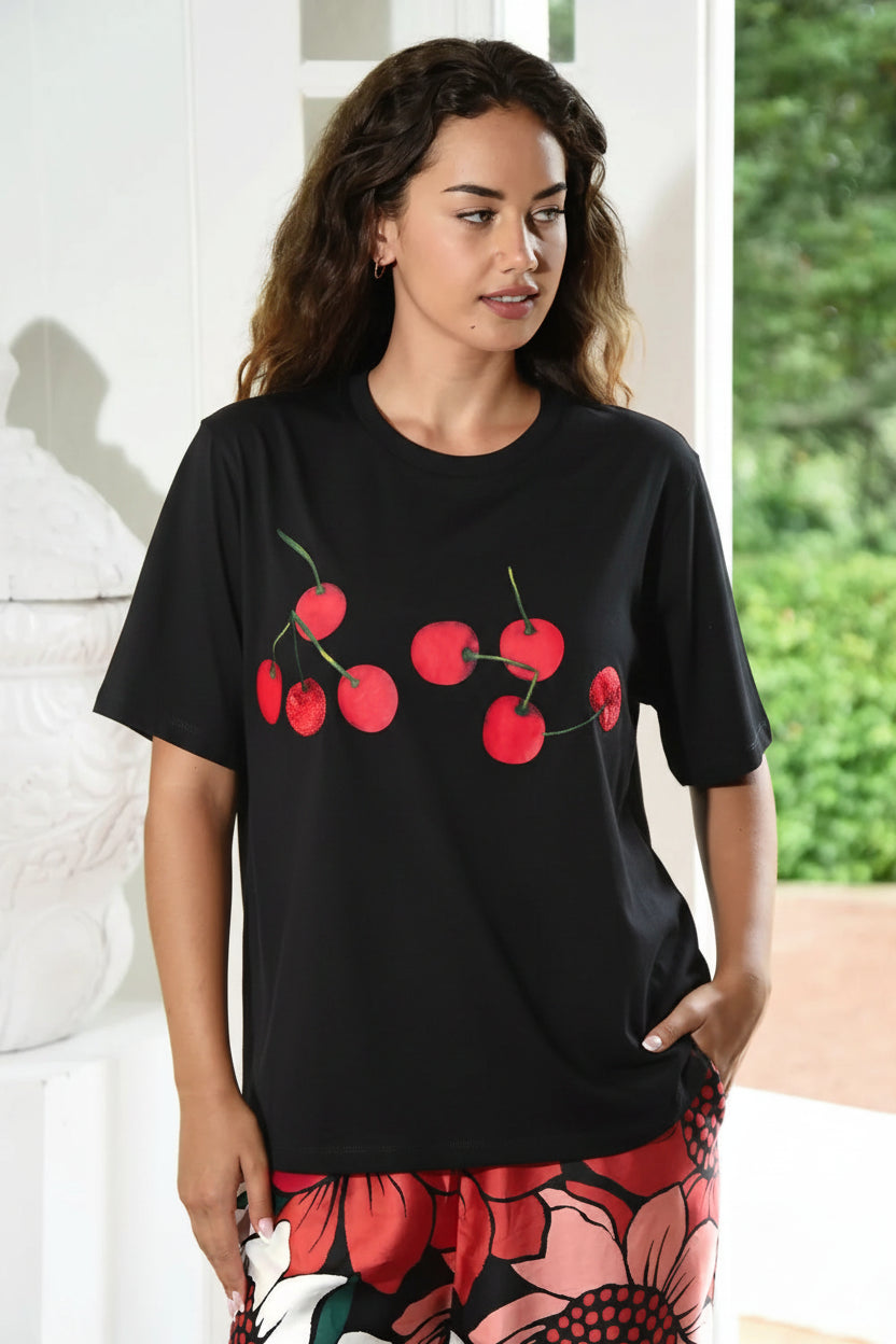 A woman wearing a coral-red short-sleeve t-shirt with a cherry graphic print on the front, paired with a floral-patterned skirt, standing in a bright indoor setting.