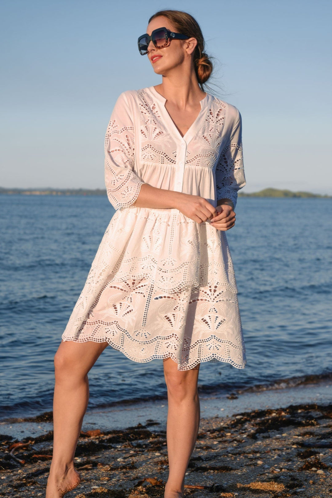 Woman in a white lace curate by trelise cooper dress standing on a beach with ocean in the background