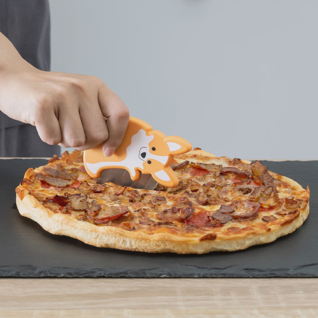 Person using a cow-shaped pizza cutter on a pizza on a black slate board.