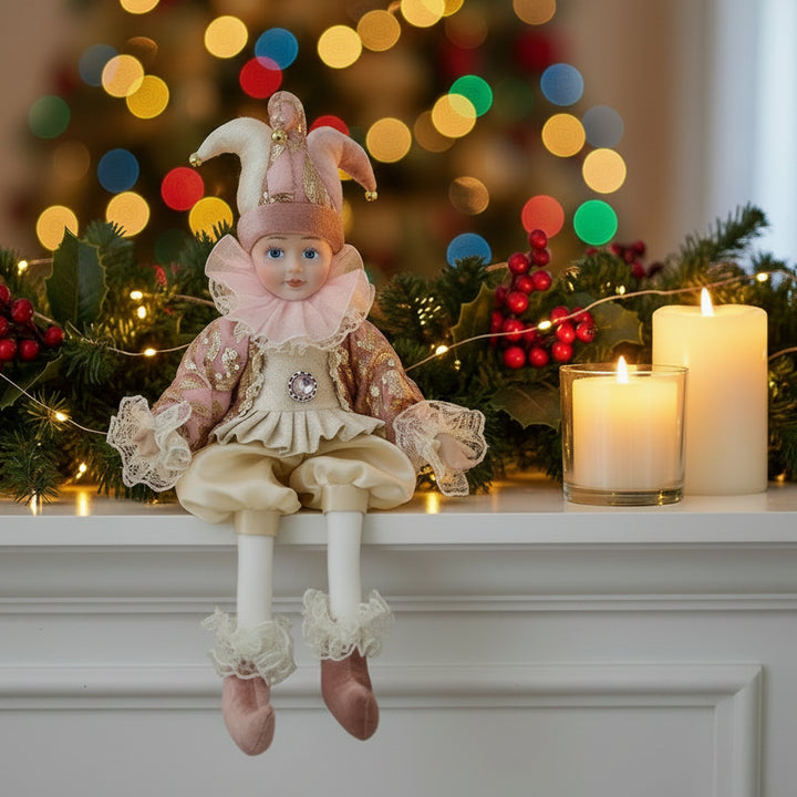 A vintage-style porcelain doll dressed as a jester, featuring a pink and gold hat, ruffled collar, patterned jacket, cream breeches, and pink shoes, isolated on a white background.