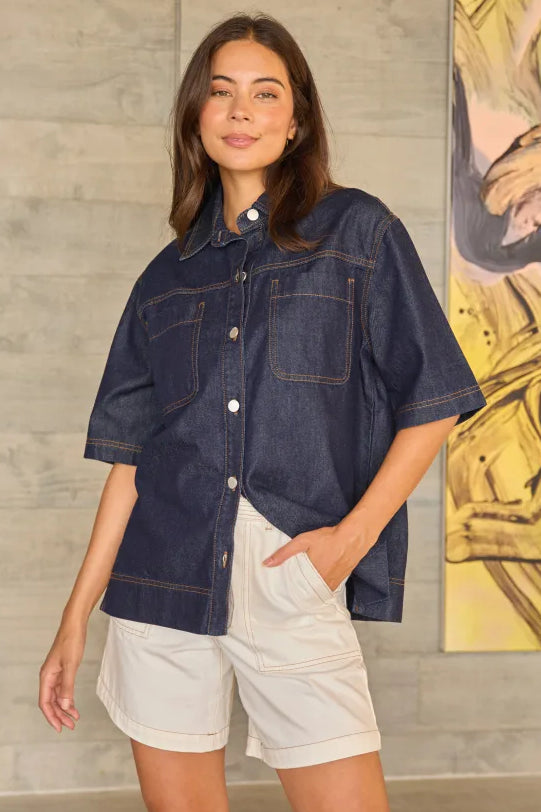 A woman modeling a dark indigo blue short-sleeve button-up denim shirt with white shorts, standing in front of a textured gray wall with a yellow abstract painting on the side.