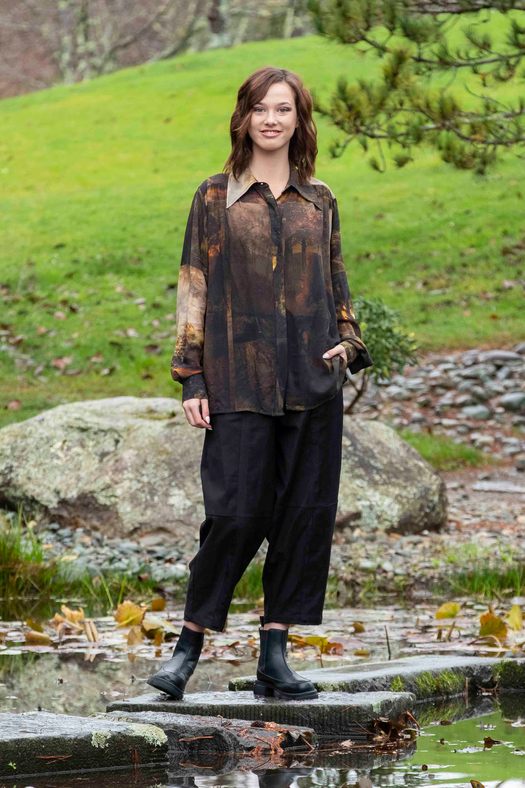 Person standing on a stone path by a pond with green grass and trees in the background